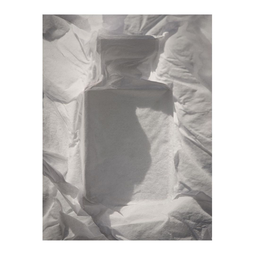 Product shot of a clear glass bottle surrounded by wrinkled white tissue paper, shot with soft, diffused lighting to minimise shadows.