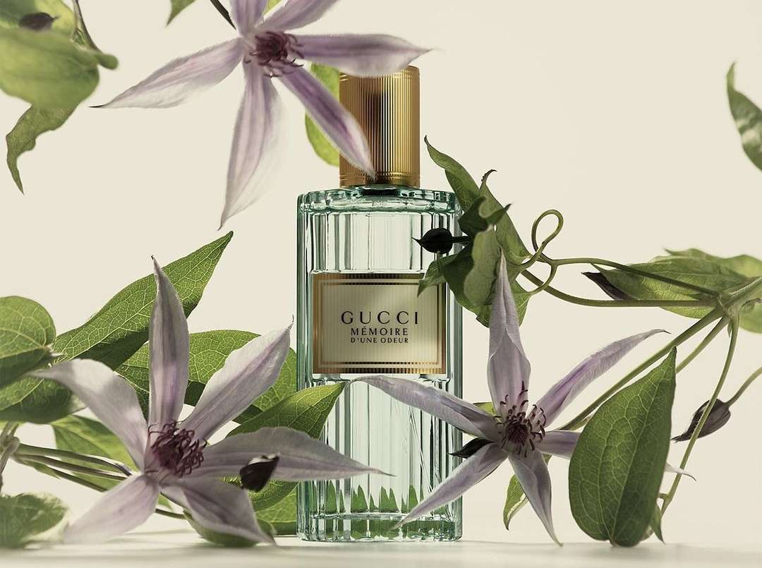 Close-up of a Gucci Mémoire d'une Odeur perfume bottle surrounded by purple clematis flowers and green leaves against a cream background.