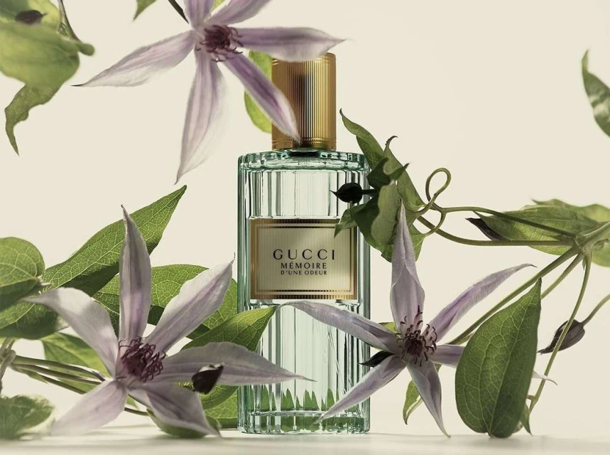 Close-up of a Gucci Mémoire d'une Odeur perfume bottle surrounded by purple clematis flowers and green leaves against a cream background.