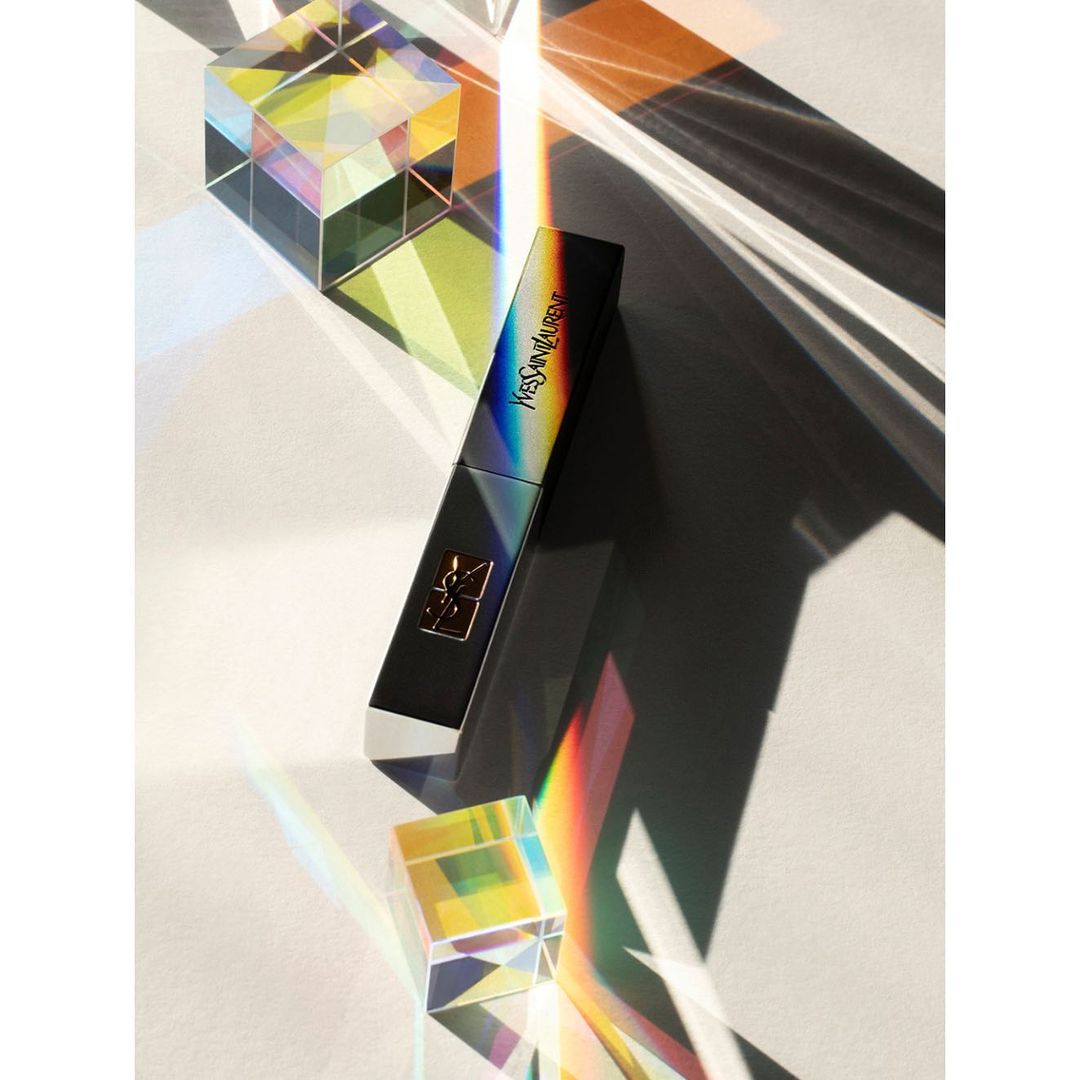 Still life featuring a YSL lipstick with holographic packaging, placed beside a prism reflecting rainbow hues on a white surface.