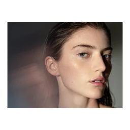 Close-up portrait of a woman with wet hair and a dewy complexion looking toward the camera against a neutral background.