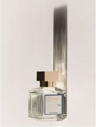 Glass perfume bottle with a square gold cap casting a tall dark shadow against a neutral textured wall background.