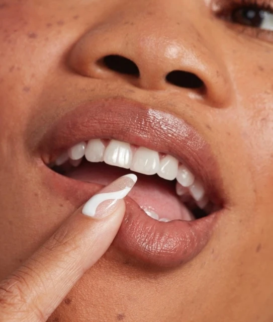 Close-up of a woman's face with white teeth, brown lips with lip gloss, and a finger with a nail design touching her lips.