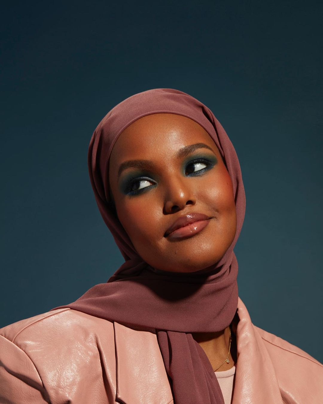 Woman with dark skin wearing a headscarf and light pink leather jacket, looking up and to the side against a teal background.