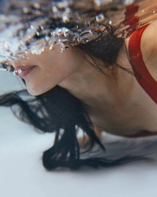 Underwater view of a woman with dark hair, wearing a red top, surrounded by clear water and bubbles, partially submerged.
