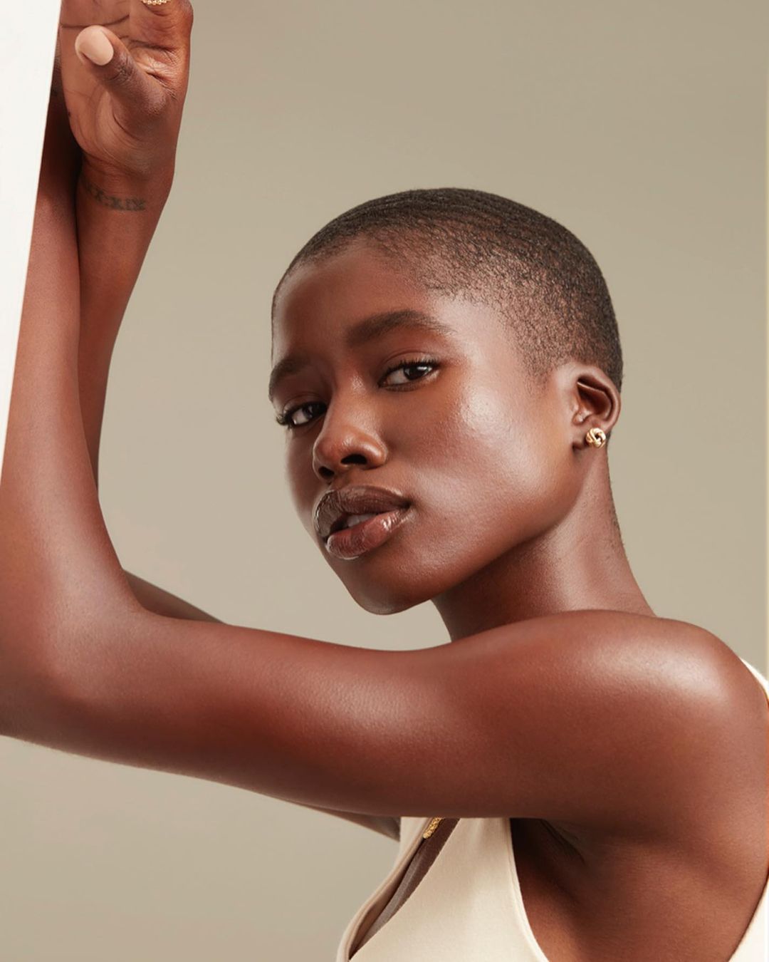 Close-up studio shot of a young Black woman with a buzz cut, wearing a white top and gold earrings, posing against a neutral backdrop.