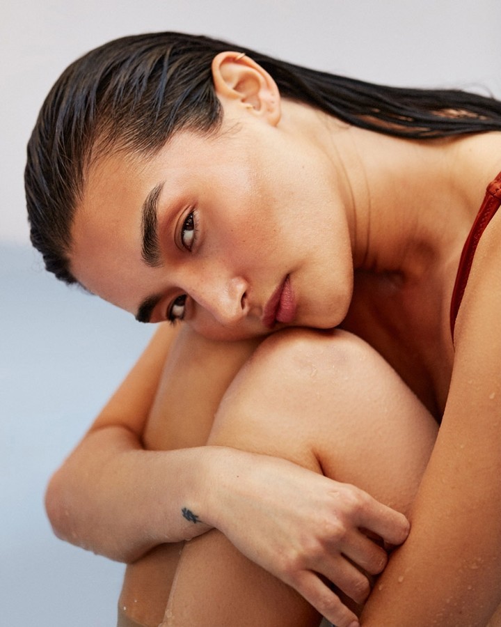 Portrait of a woman with wet hair, embracing her knees in a light blue environment, creating a serene and intimate atmosphere.