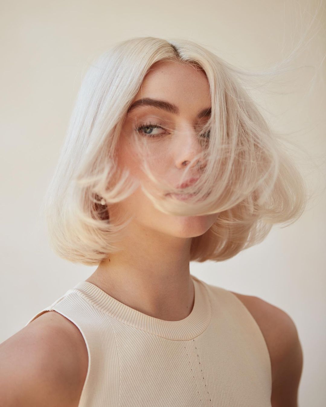 Close-up portrait of a young adult woman with wavy platinum blonde hair and a light complexion, wearing a sleeveless top, against a soft background.