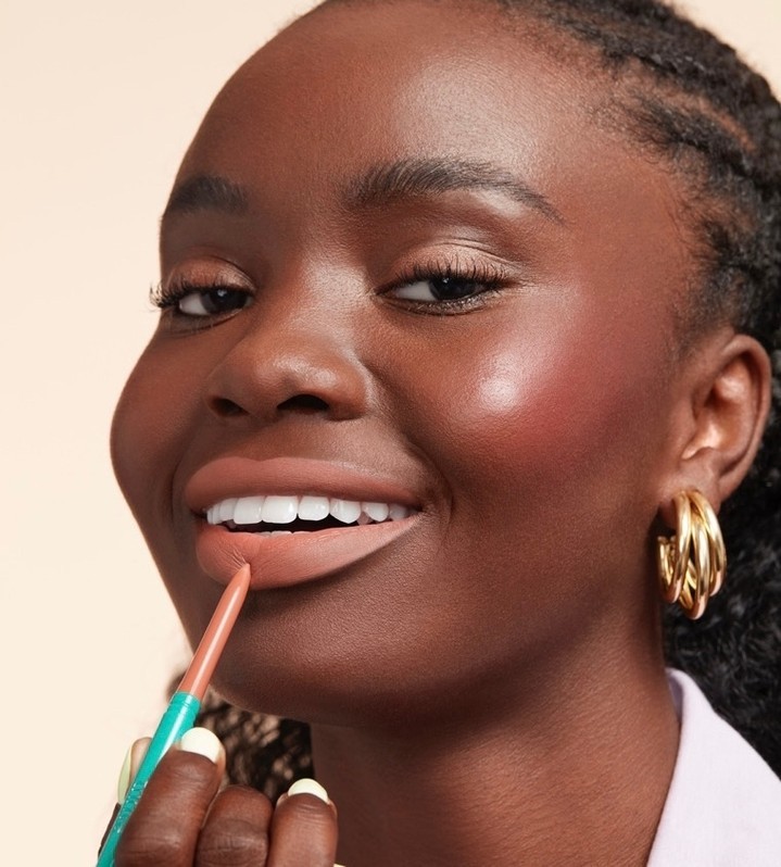 Close-up of a woman applying nude lip liner. She has dark skin, full lips, gold hoop earrings, and wears a light colored top.