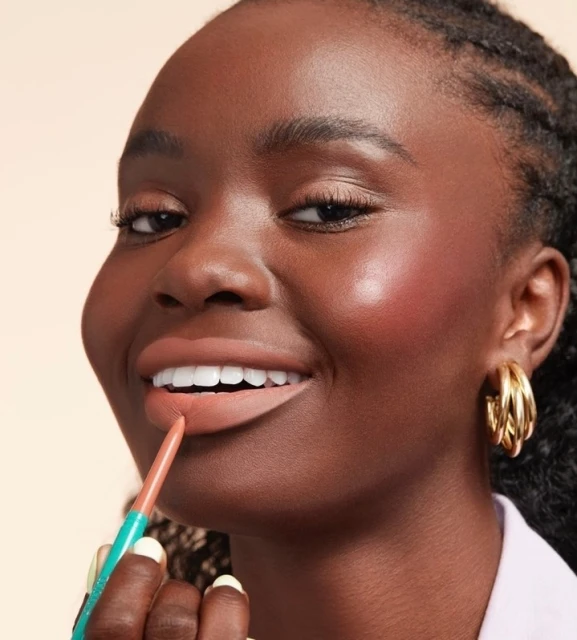 Close-up of a woman applying nude lip liner. She has dark skin, full lips, gold hoop earrings, and wears a light colored top.