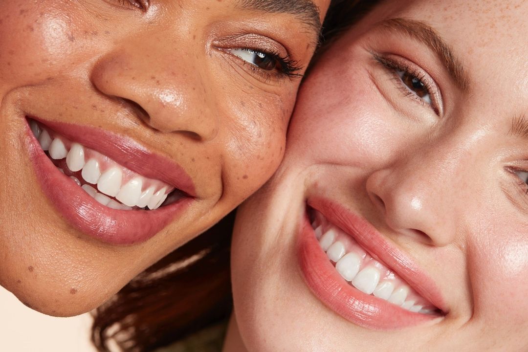 Close-up shot of two smiling women with flawless skin and pink lip gloss, demonstrating a beauty or skincare product for advertising.