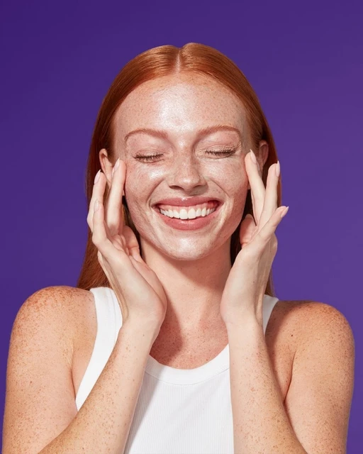 Close-up of a red-haired woman with freckles, smiling with her eyes closed, holding her hands up to her face against a purple background.
