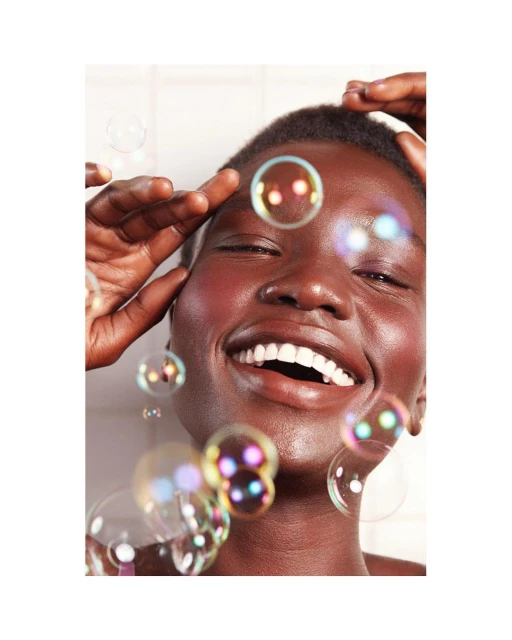 Close-up of a smiling Black woman surrounded by floating iridescent soap bubbles. She is touching her forehead, and the background is white.