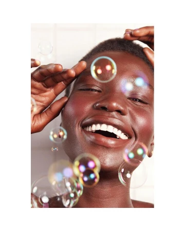 Close-up of a smiling Black woman surrounded by floating iridescent soap bubbles. She is touching her forehead, and the background is white.