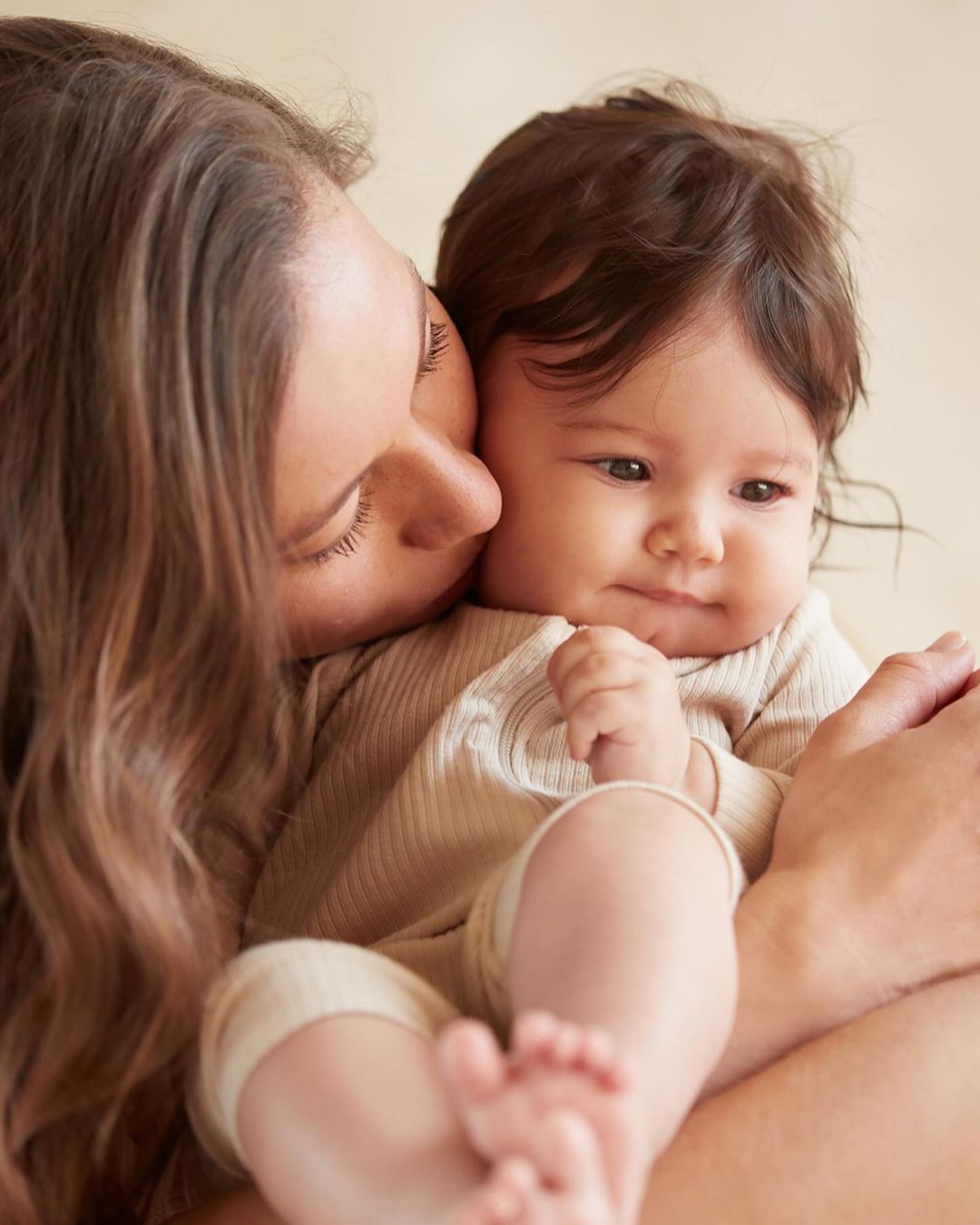 A close-up shot shows a young mother with long brown hair gently kissing her baby's cheek, both dressed in neutral-toned clothing.