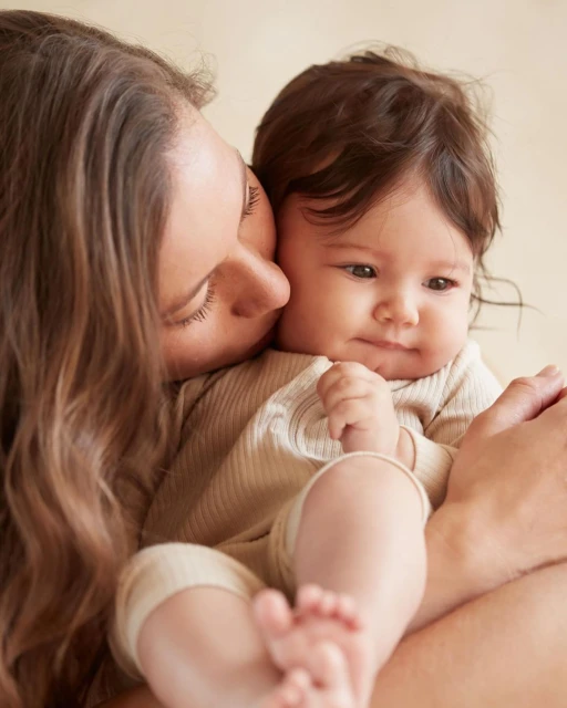 A close-up shot shows a young mother with long brown hair gently kissing her baby's cheek, both dressed in neutral-toned clothing.