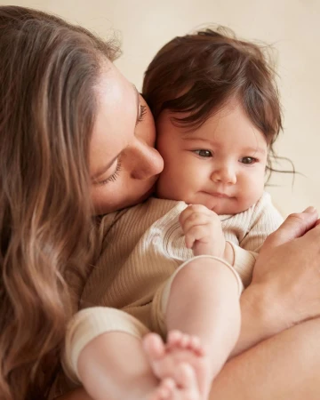 A close-up shot shows a young mother with long brown hair gently kissing her baby's cheek, both dressed in neutral-toned clothing.