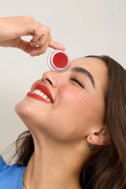 Smiling woman with closed eyes and red lipstick posing with a jar of red cream, showcasing skincare and beauty in a close-up studio shot.