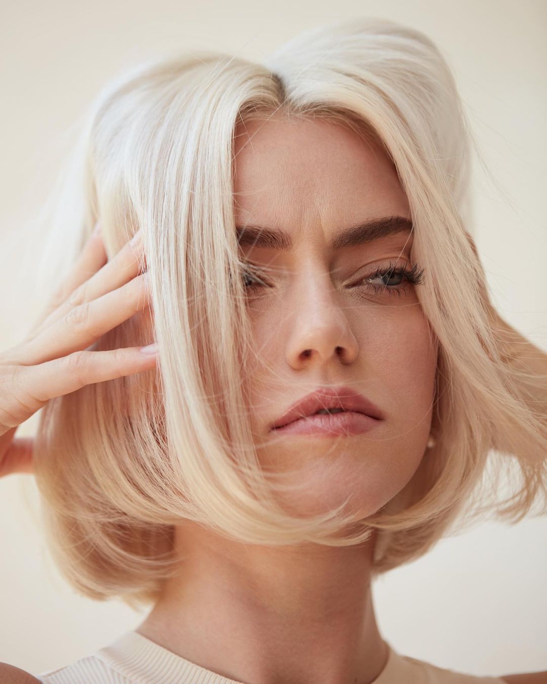 Close-up portrait of a young adult woman with light blonde bob haircut, posing with hands near her face in a studio setting.