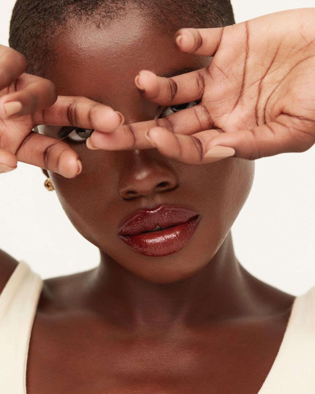 Close-up portrait of a dark-skinned woman partially covering her face with her fingers, featuring glossy lips and a neutral background.