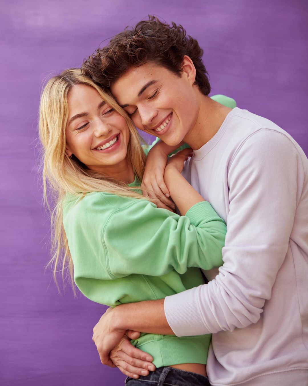 Smiling couple embracing in front of a solid purple backdrop. The woman has blonde hair, and the man has brown hair. They wear casual sweaters.