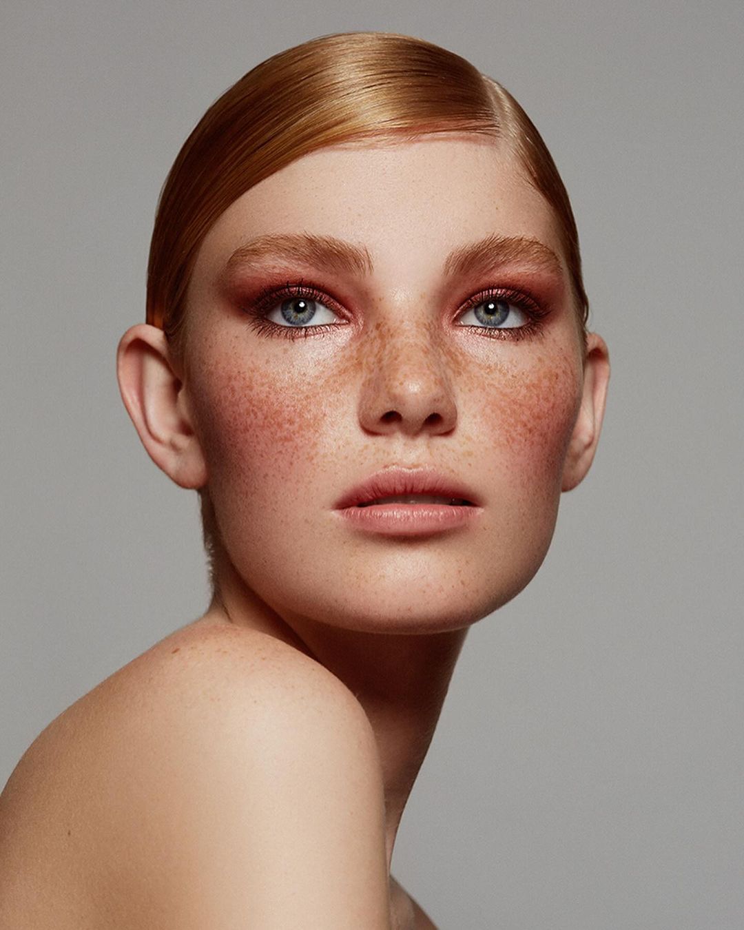 Close-up portrait of a young adult woman with red hair and freckles, wearing makeup and posing in front of a neutral gray backdrop.