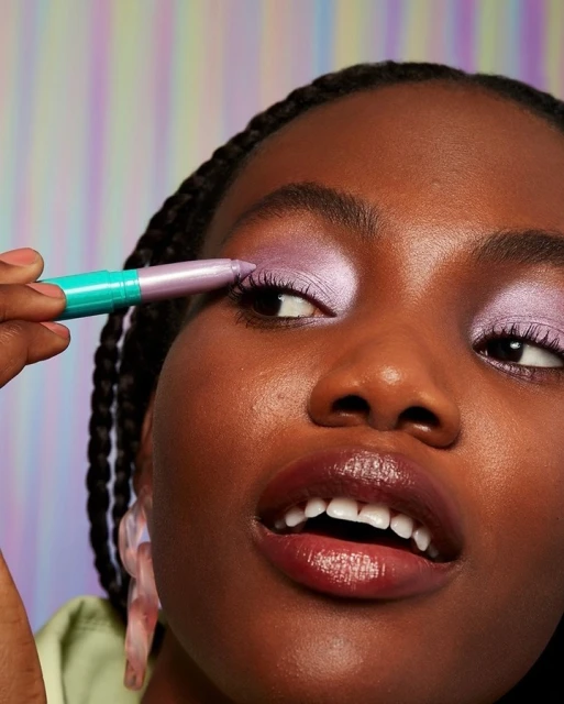 Close-up of a young Black woman applying lavender eyeshadow with a turquoise pencil. She is smiling with a rainbow background.