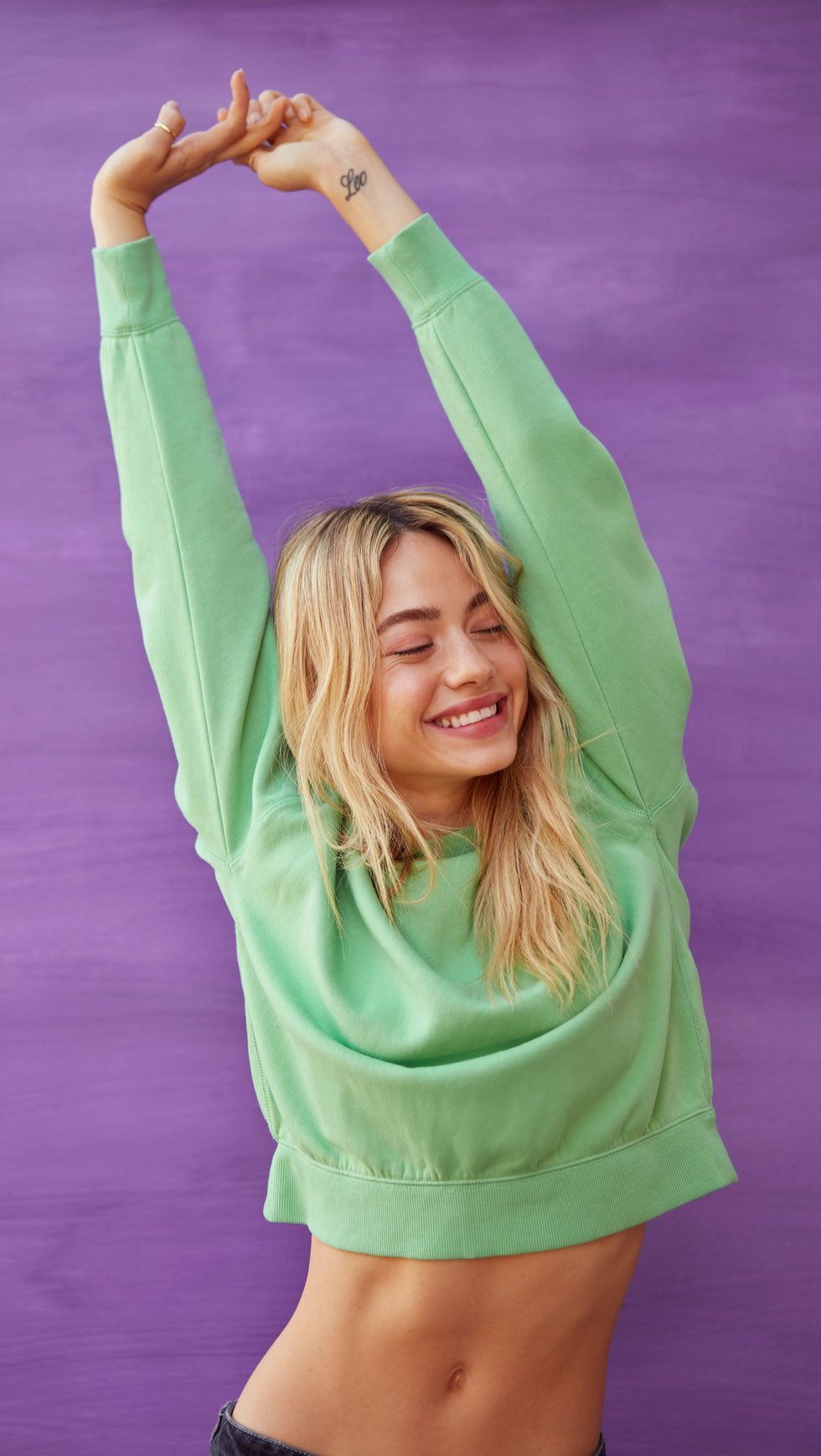 Smiling adult woman in a green sweatshirt stretching with her arms overhead against a purple background in a studio shot.