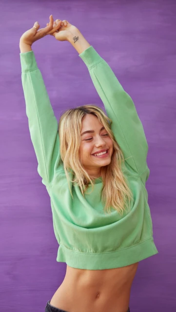 Smiling adult woman in a green sweatshirt stretching with her arms overhead against a purple background in a studio shot.