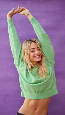 Smiling adult woman in a green sweatshirt stretching with her arms overhead against a purple background in a studio shot.
