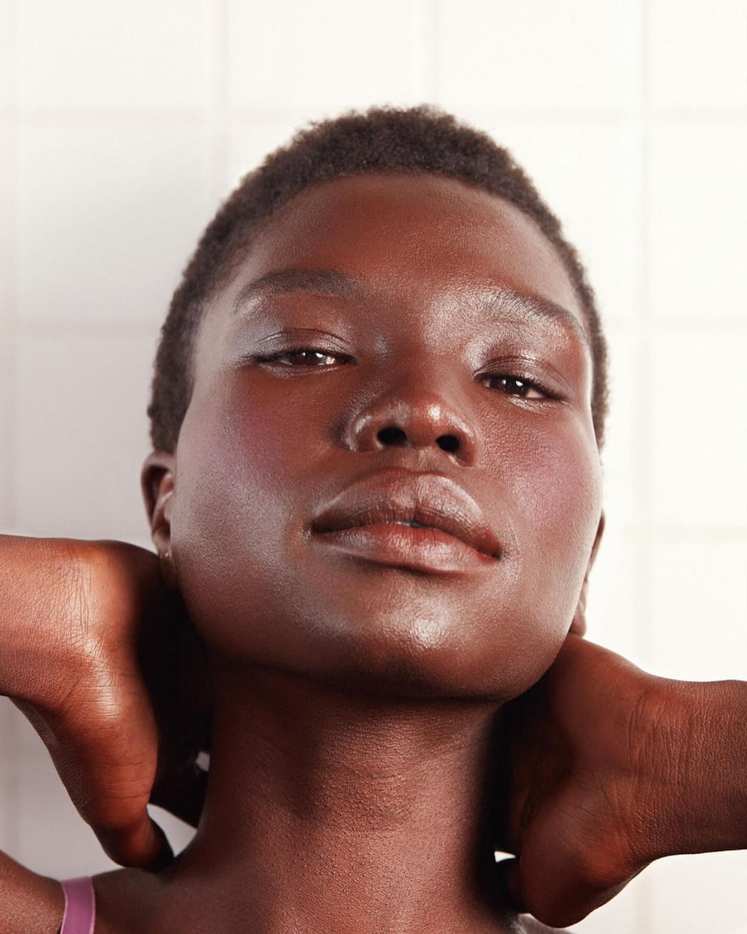 Close-up studio portrait of a dark-skinned woman with hands behind her head, featuring minimal makeup and a white tiled background.