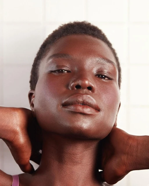 Close-up studio portrait of a dark-skinned woman with hands behind her head, featuring minimal makeup and a white tiled background.
