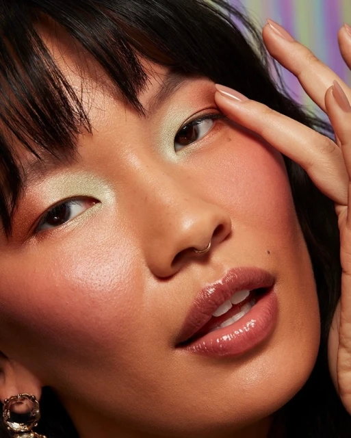Close-up of a woman with bangs and a nose ring, applying light green eyeshadow with her finger, and wearing shimmery peach blush.
