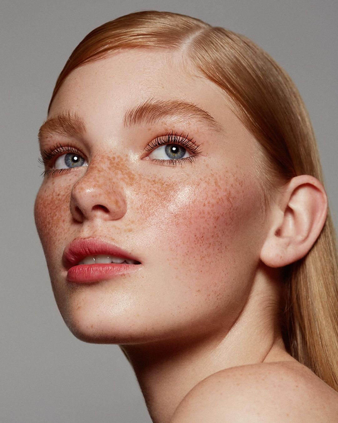 Close-up portrait of a young adult woman with freckles, fair skin, light-red hair, and light-pink lips against a gray background in a photo studio.