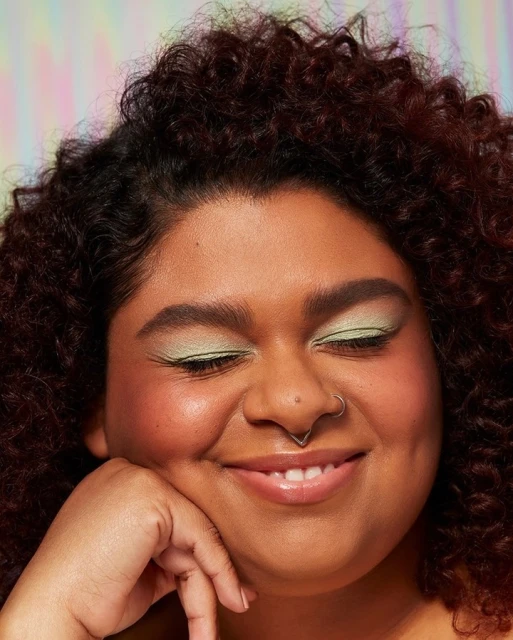 Close-up shot of a woman with curly hair and green eye shadow smiling with her eyes closed against a vibrant background.