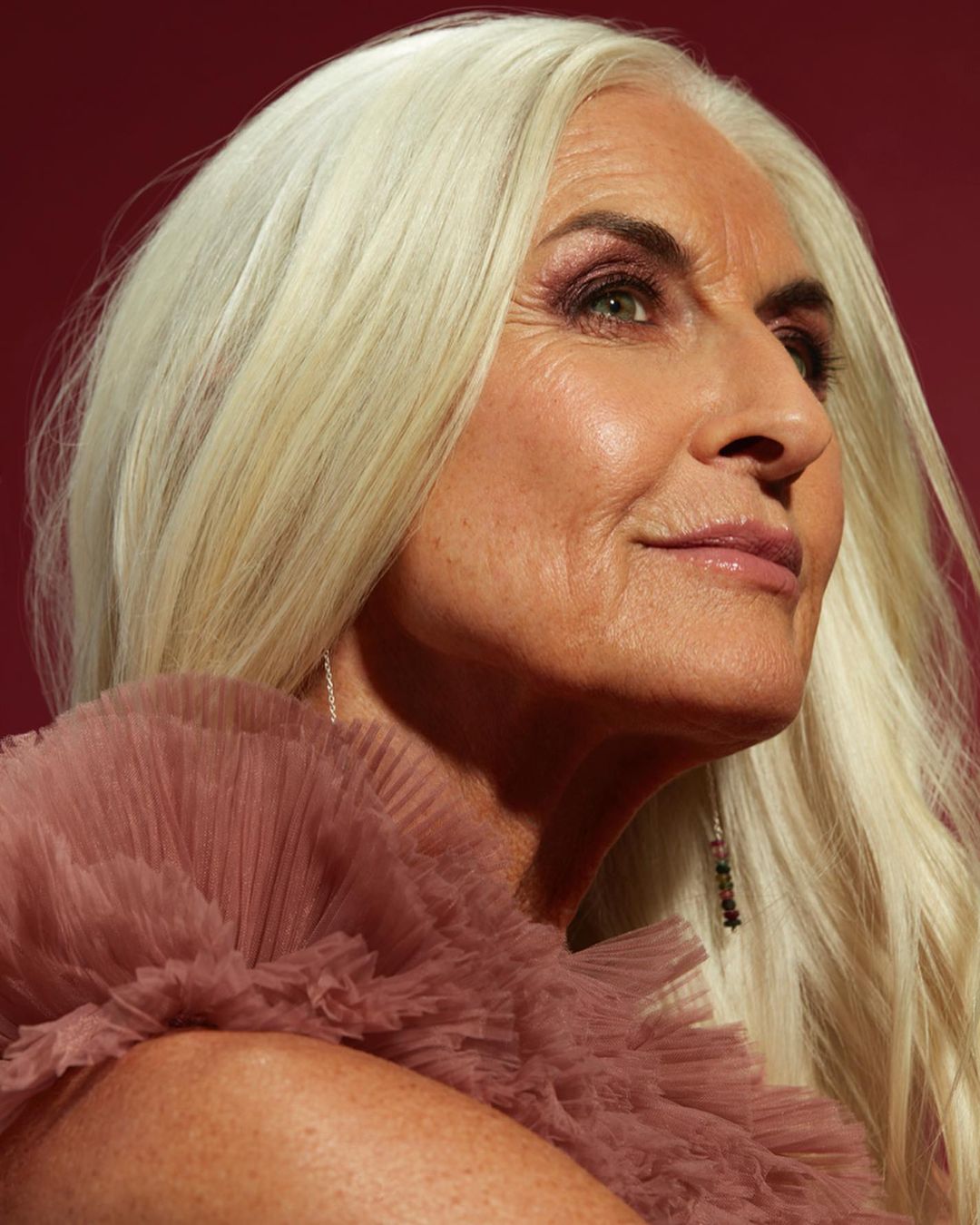 Close-up portrait of an adult woman with long white hair wearing a frilly, textured mauve top against a maroon background.