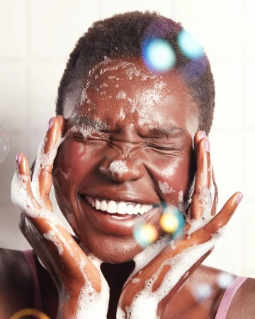 Close-up of a Black woman washing her face with soapy lather, smiling with eyes closed, captured by Beth Sternbaum.