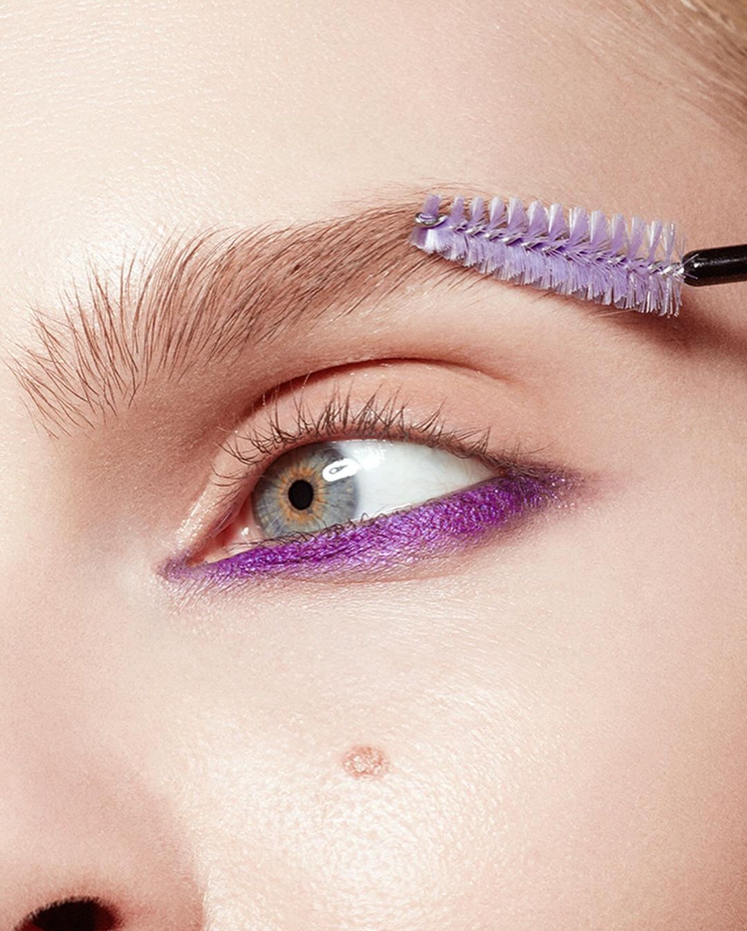 Close-up shot of a woman's face with light skin and an eyebrow brush applying make-up. Purple eyeliner is visible, and her eye is gray.