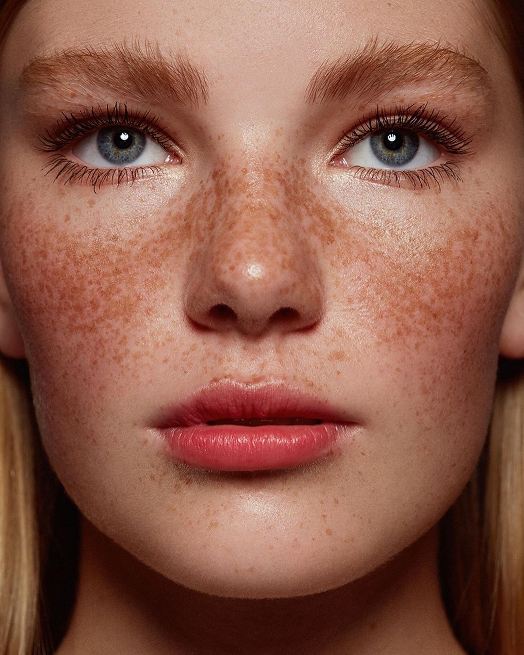 Close-up portrait of a young woman with freckles and blue eyes, showcasing her natural beauty in soft lighting, suitable for beauty or fashion themes.