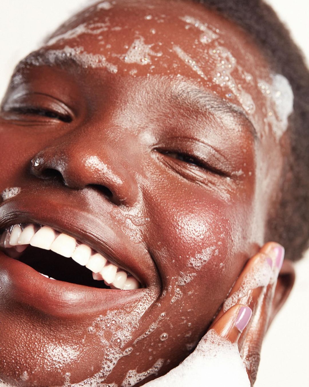 Close-up of an African-descent woman smiling with soapy cleanser on her face, showcasing radiant skin and joyful expression against a light background.