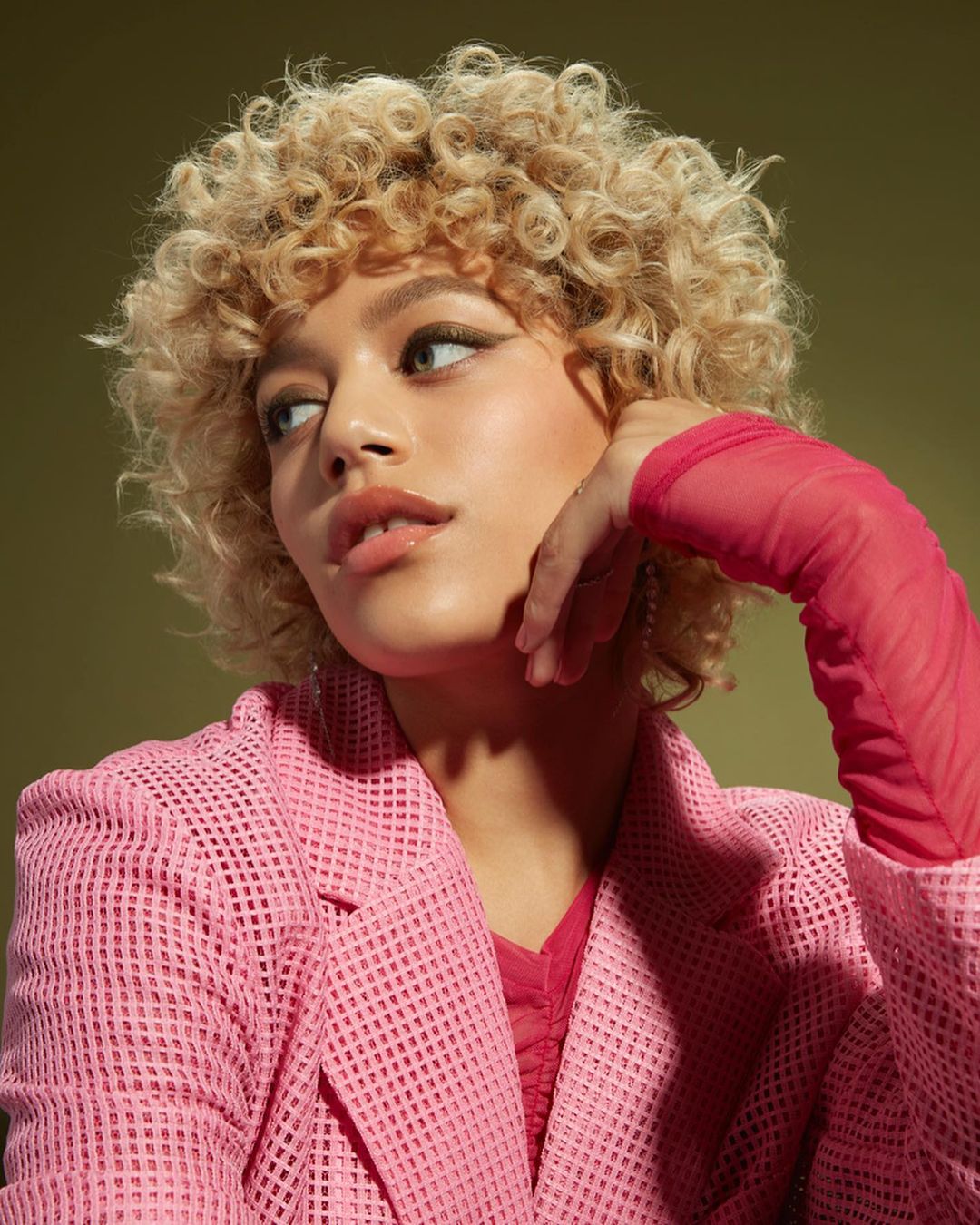 Close-up studio portrait of a woman with curly blonde hair wearing a pink mesh jacket, set against a solid olive green backdrop.