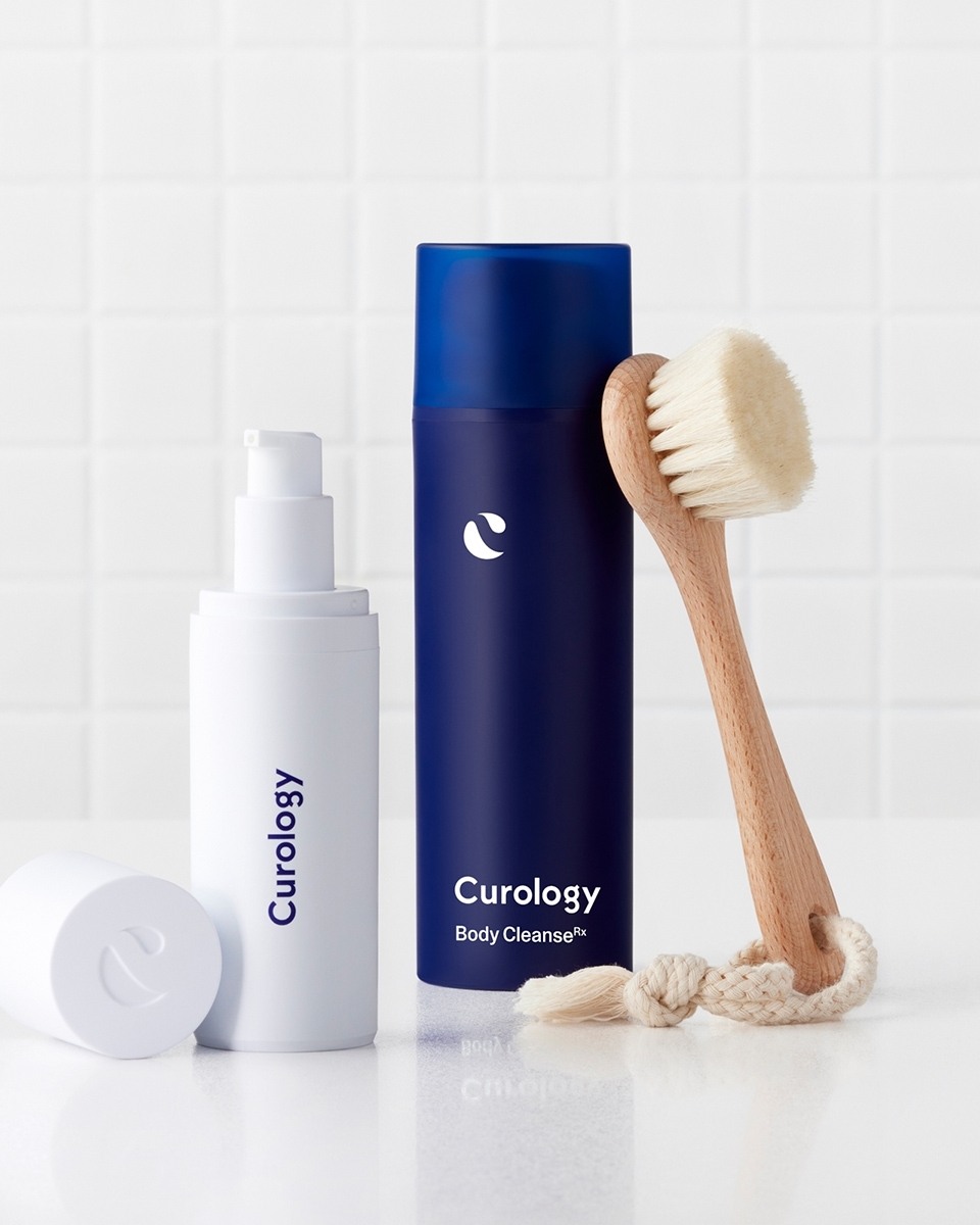 Curology skincare products: cleanser and body cleanser displayed with a facial brush on a white surface against a white tiled background.