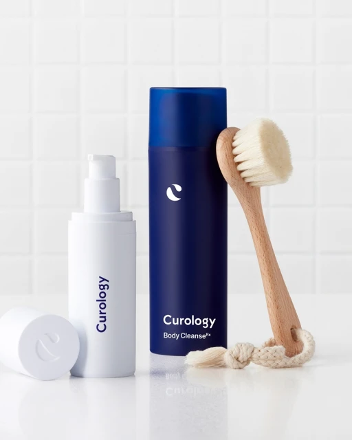 Curology skincare products: cleanser and body cleanser displayed with a facial brush on a white surface against a white tiled background.