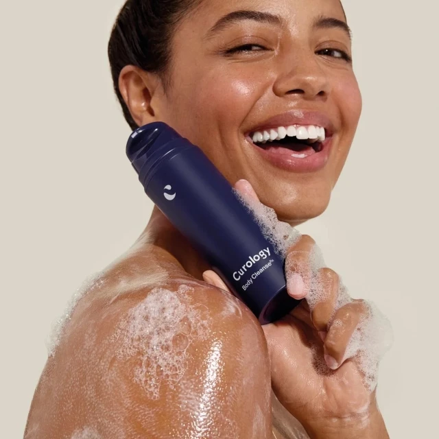 Smiling woman holds a blue Curology cleanser, with foam on her skin, set against a neutral background in a beauty product shot.