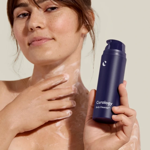 An adult woman holds a bottle of Curology Body Cleanse with white cleansing foam on her chest and neck against a neutral background.