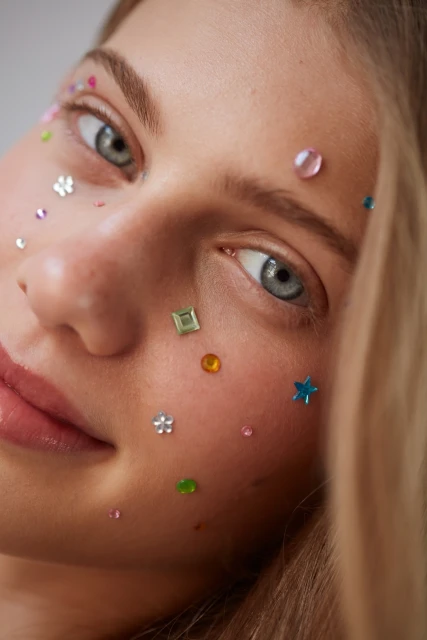 Close-up of a young, fair-skinned woman with blue eyes and blonde hair. She has colorful gem-like makeup on her face in studio lighting.