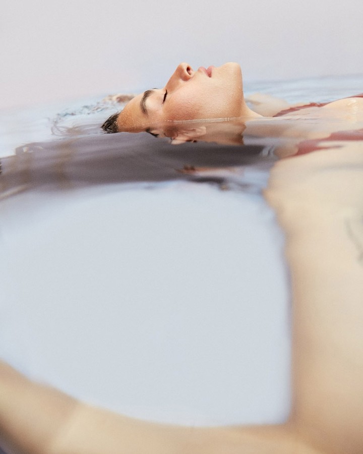Adult female floats in calm water, eyes closed in relaxation, indoor setting, light complexion, fair lighting, horizontal orientation.