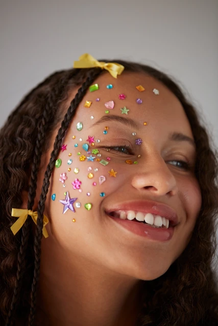 Close-up portrait of a smiling young woman with braided hair adorned with colorful star and jewel-shaped face gems against a neutral backdrop.