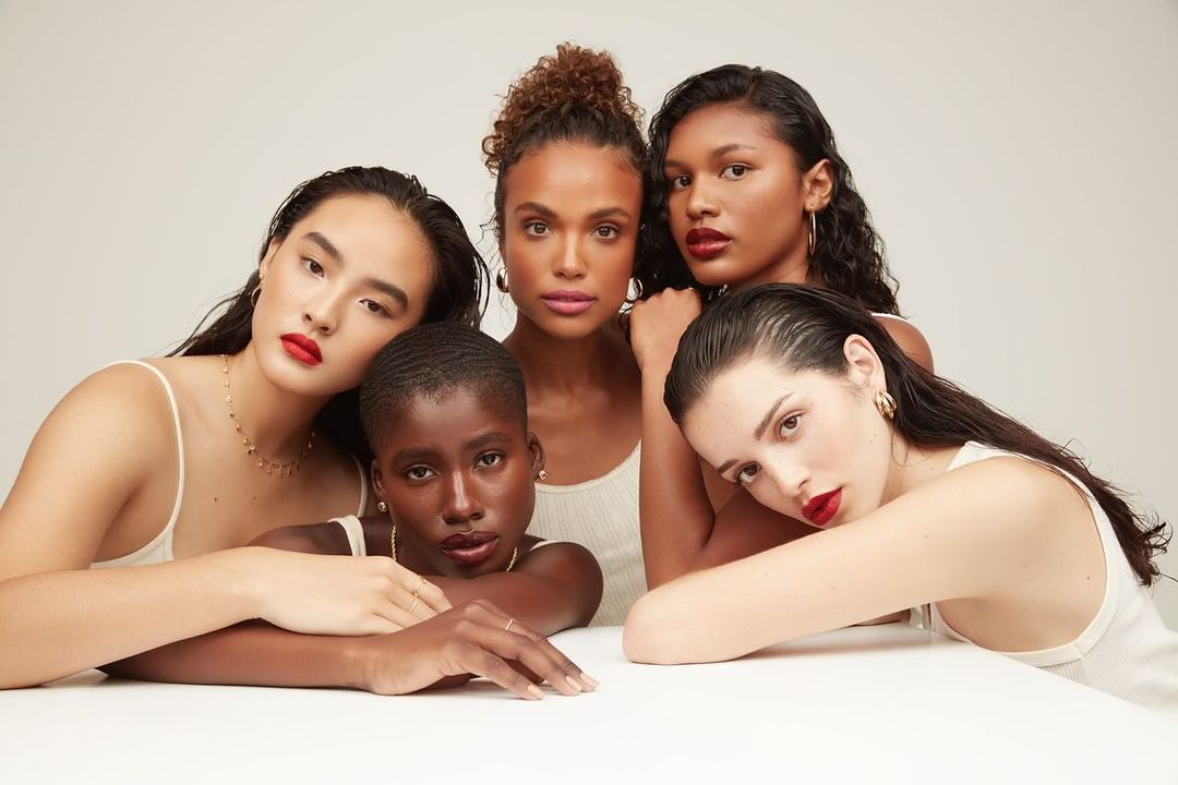 Group of five diverse women with red lipstick posing in front of a neutral background, wearing neutral tops, and gold jewelry.