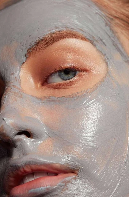 Close-up of a fair-skinned woman's face covered in a gray facial mask, highlighting her eye, skin texture, and beauty treatment.